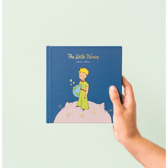 Main plaçant une photo sur une page autocollante de l'album Le Petit Prince