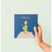 Main plaçant une photo sur une page autocollante de l'album Le Petit Prince